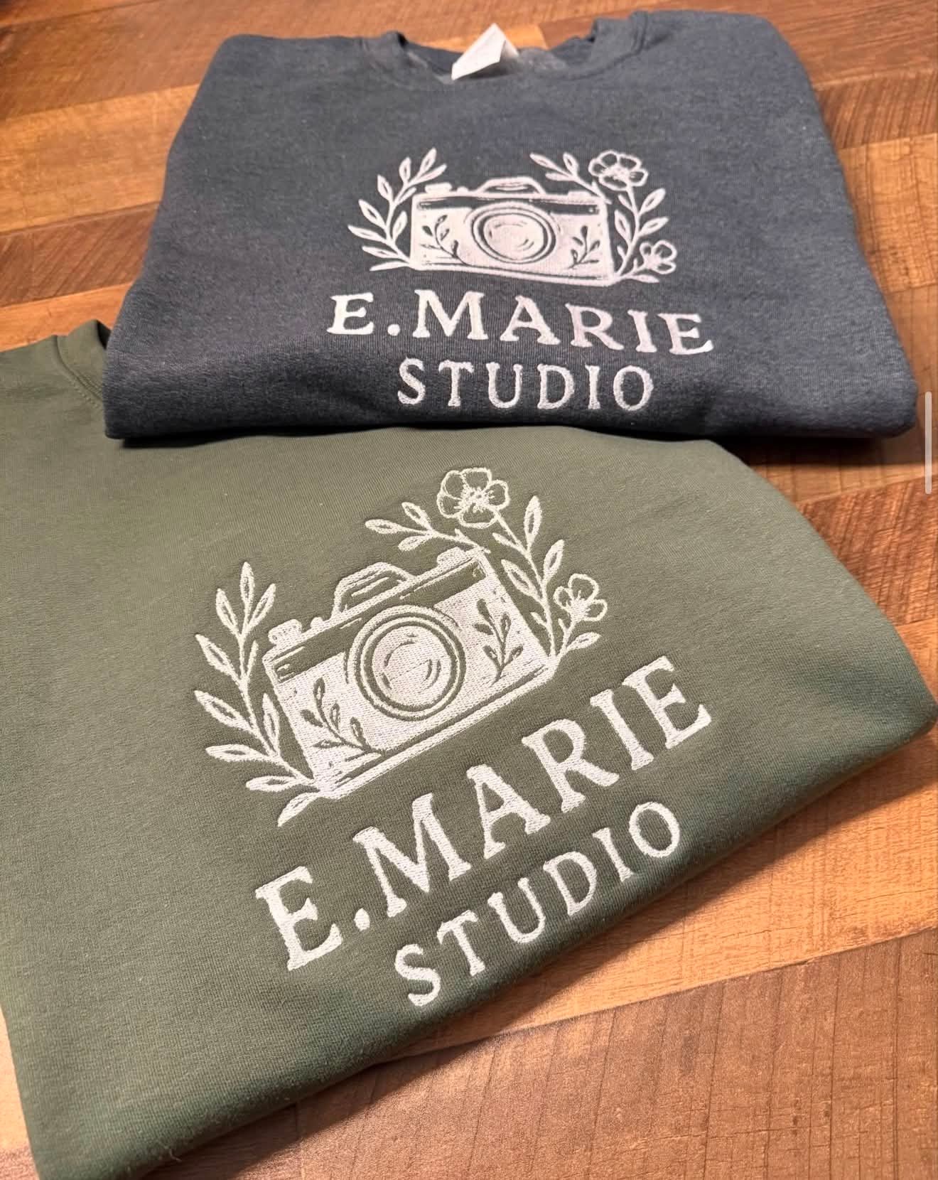 Navy and olive embroidered crewnecks with a camera and floral design reading E. Marie Studio