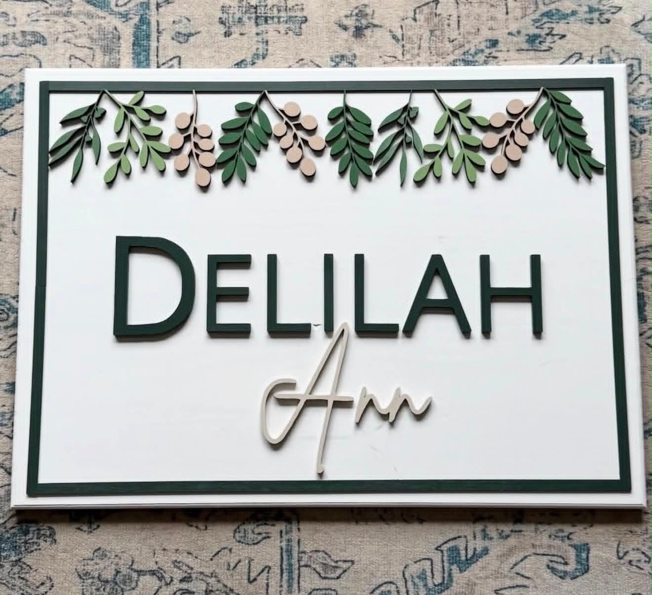 White nursery sign reading Delilah Ann with a hand-painted floral garland along the top