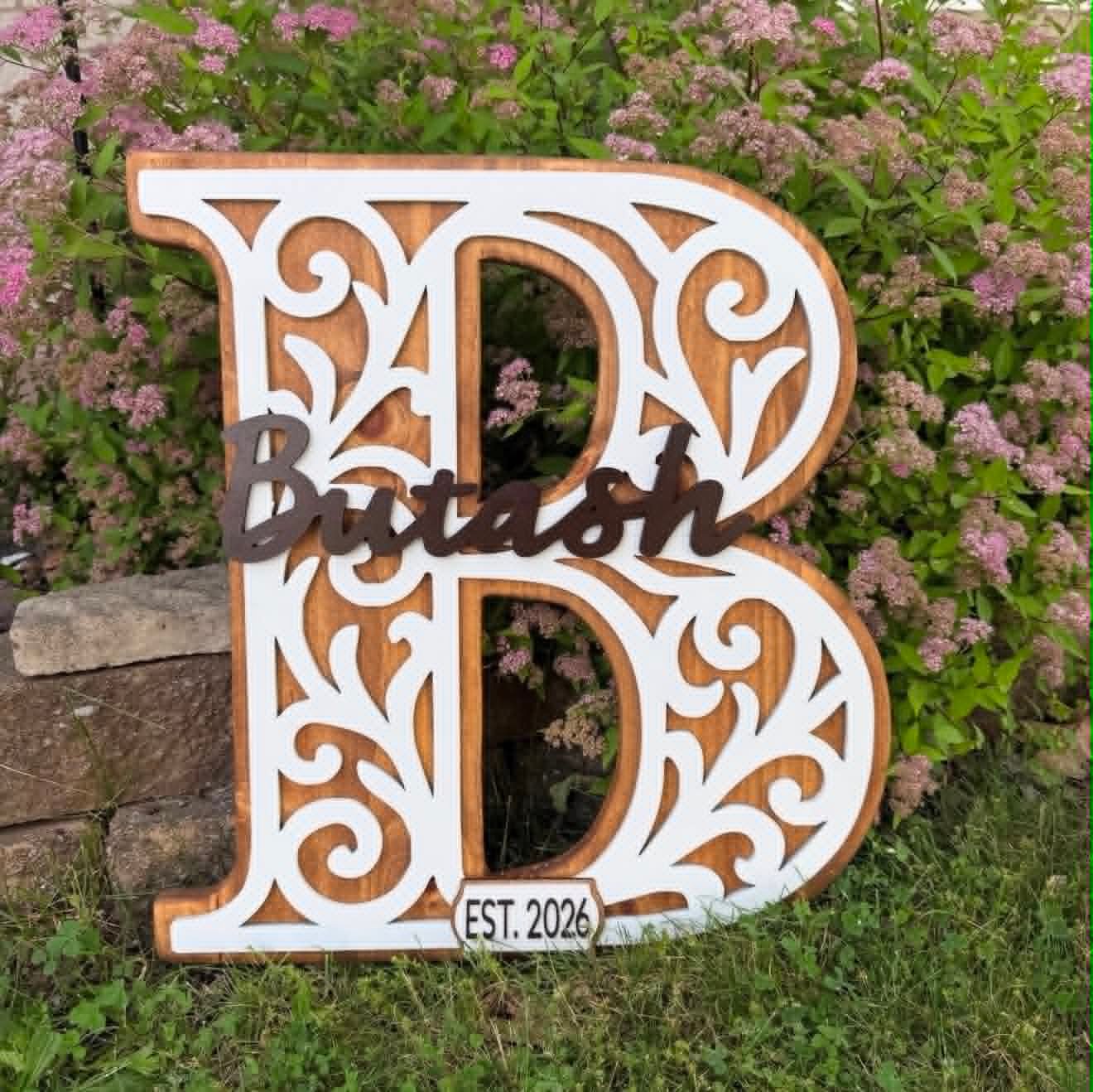 Large monogram letter B sign with white scrollwork over stained wood, reading Butash Est. 2026